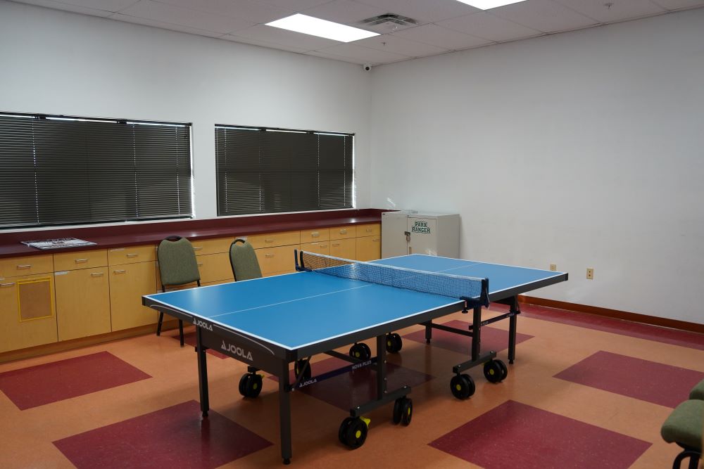 Bright activity room with a blue ping pong table centered in front of cabinets and windows